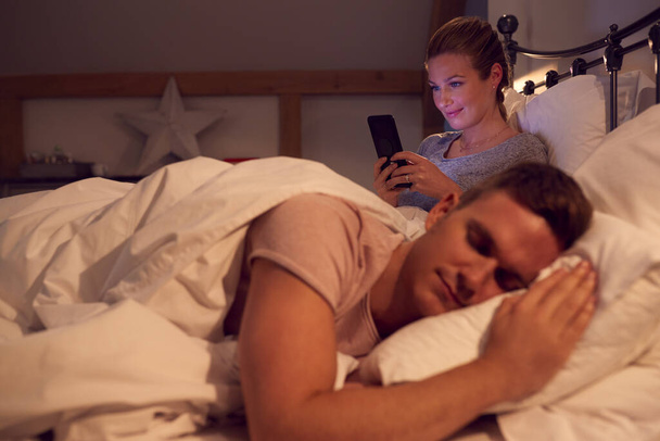 Woman At Home Using Mobile Phone In Bed Whilst Man Sleeps - Foto, afbeelding
