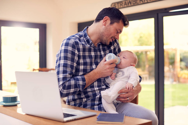 Working Father Using Laptop At Home Whilst Feeding Baby Son On Knee With Bottle - Zdjęcie, obraz