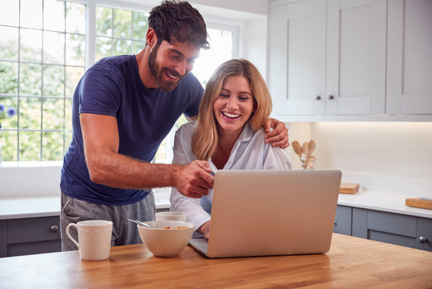 Couple Wearing Pyjamas In Kitchen Watching Laptop Whilst Eating Breakfast - Photo, image