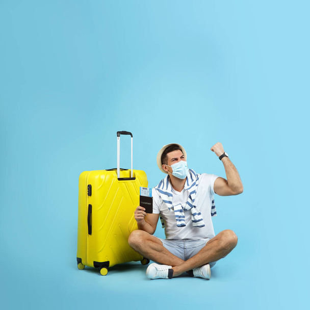 Male tourist in protective mask holding passport with ticket and suitcase on turquoise background, space for text - Фото, зображення