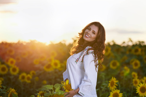 Beautiful woman enjoying nature in the sunflower field at sunset. Traditional clothes. Attractive brunette woman with long and healthy hair. - Zdjęcie, obraz