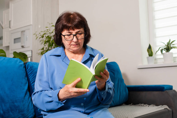 Senior woman reading, sitting on blue sofa - Fotó, kép