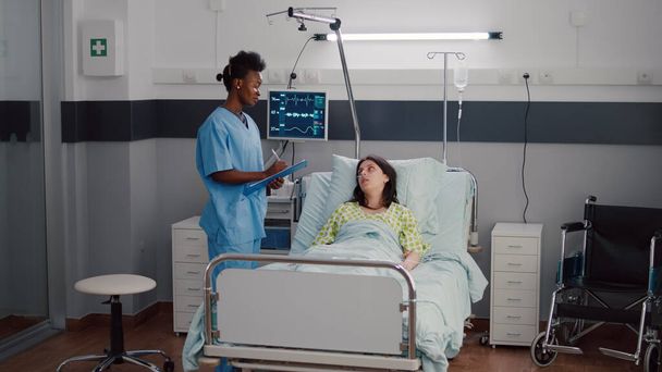 Primer plano de enfermera afroamericana monitoreando a mujer enferma mientras descansa en la cama - Foto, Imagen