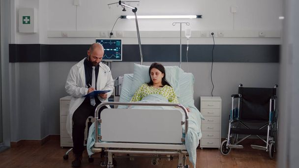 Vista frontal de una mujer enferma descansando en la cama discutiendo con un oncólogo médico - Foto, imagen
