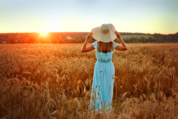 Piękna młoda kobieta ubrana w niebieską sukienkę i kapelusz cieszy się na świeżym powietrzu, patrząc na słońce na idealnym polu pszenicy o zachodzie słońca - Zdjęcie, obraz