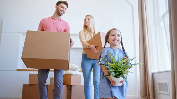Funny little daughter helps parents with brown cardboard packages move holding small green pot plant in new flat - Foto, imagen