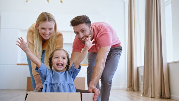 Смішні батьки леді блондинка і бородатий хлопець переміщують велику картонну коробку з в захваті дошкільнята дівчина
 - Фото, зображення