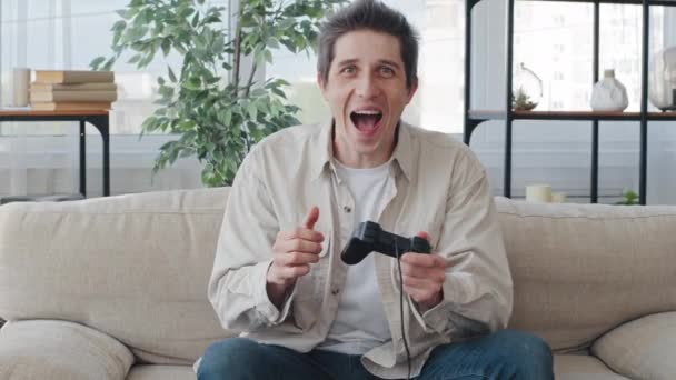 Caucasian man male millennial guy gamer sitting at home on couch playing video game console using controller joystick enjoying gaming shouts with joy celebrating winning making victory hand gesture - Footage, Video