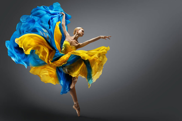 Beautiful Woman Ballet Dancer Jumping in Air in Colorful Fluttering Dress. Graceful Ballerina Dancing in Yellow Blue Gown over Gray Studio Background - Фото, изображение