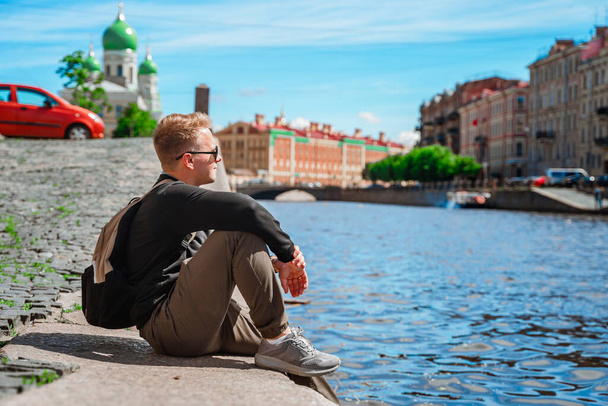  橋や運河の美しい景色を望むサンクトペテルブルクの堤防に沿って若い男が歩く. - 写真・画像
