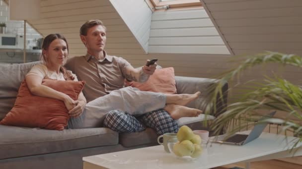 Követés balra boldog heteroszexuális kaukázusi pár ül a kanapén otthon, TV-t néz, férfi használja távirányító és nő beszél és mosolyog - Felvétel, videó