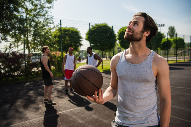 Sportsman with basketball ball looking away near blurred interracial friends on playground  - Foto, Imagen