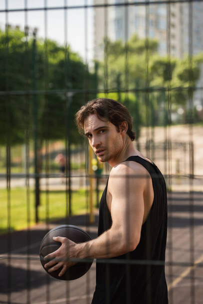 Young man with basketball ball looking at camera near fence outdoors  - Photo, Image