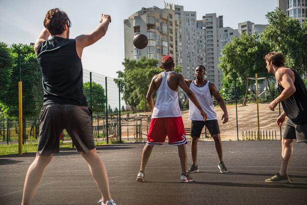 Interracial men playing basketball near friend with raised hands on blurred foreground  - Fotografie, Obrázek
