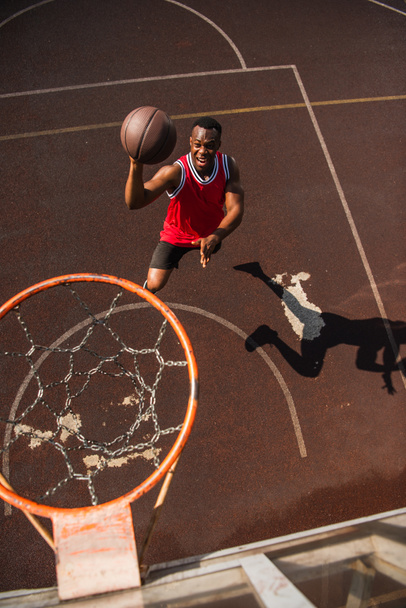 High angle view of excited african american sportsman jumping with basketball ball near hoop  - Fotó, kép