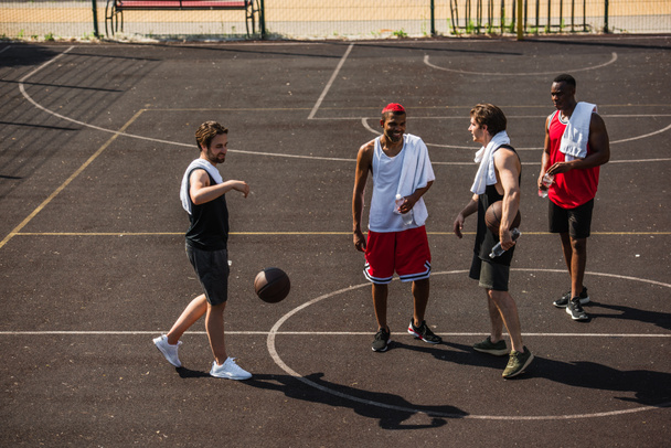 Smiling multiethnic sportsmen with basketball balls and towels talking on playground outdoors  - Fotó, kép