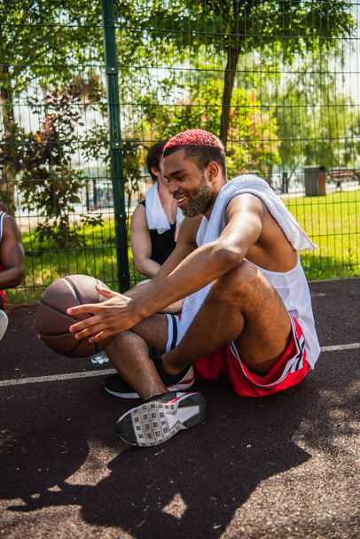 Sonriendo hombre afroamericano sosteniendo pelota de baloncesto cerca de amigos en el patio de recreo  - Foto, Imagen