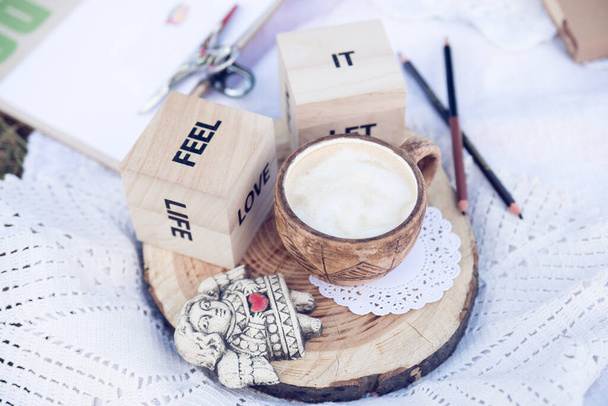 Diseño de bloques de madera con palabras "sentir", "él", "vida", "amor", pieza de madera, dos lápices, un ángel y una taza de café en ganchillo blanco con un poco de hierba en el borde. Concepto de inspiración matutina de verano. - Foto, Imagen