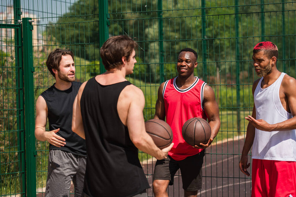Joyeux homme afro-américain tenant ballon de basket près d'amis parlant à l'extérieur  - Photo, image