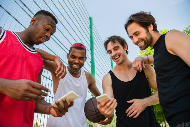 Low angle view van positieve interraciale basketbalspelers met behulp van smartphones  - Foto, afbeelding