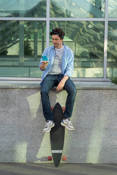 スマートフォンを使用してヒップスタースケーター男は携帯電話でオンラインでメッセージを手で読む中でロングボードを保持 - 写真・画像