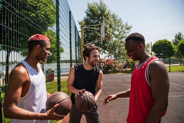Усміхнений чоловік тримає баскетбол біля афроамериканських друзів на дитячому майданчику
  - Фото, зображення