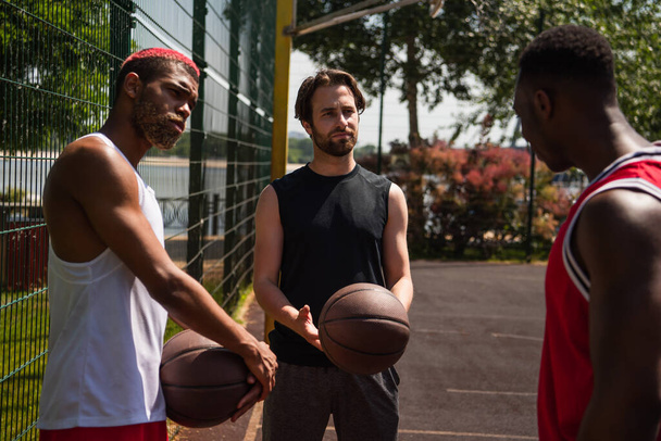 Jovens desportistas com bolas de basquete olhando para amigo afro-americano no playground  - Foto, Imagem