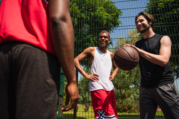 バスケットボールボールを持つ正のスポーツマンの低角度ビューでアフリカ系アメリカ人の友人を見ています。  - 写真・画像