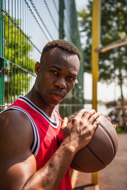 African american sportsman with basketball ball looking at camera near fence  - Foto, afbeelding