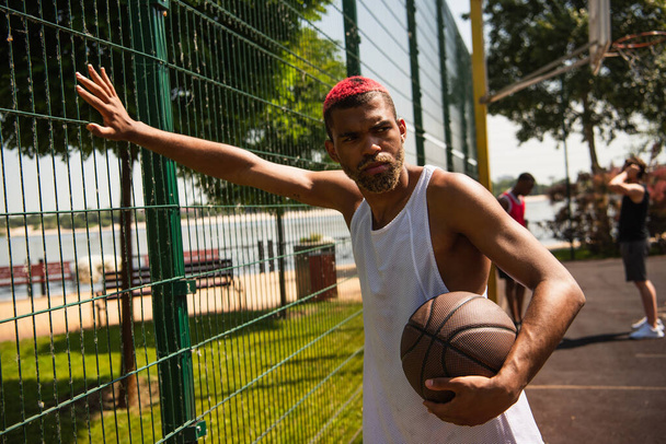 African american sportsman with dyed hair holding basketball ball near fence on court  - Фото, зображення