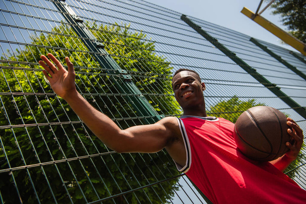 Low angle view of positive african american sportsman holding basketball ball and waving hand near fence  - Photo, Image