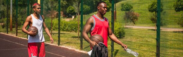 バスケットボールボールと遊び場、バナーの上を歩く水を持つアフリカ系アメリカ人スポーツマン  - 写真・画像