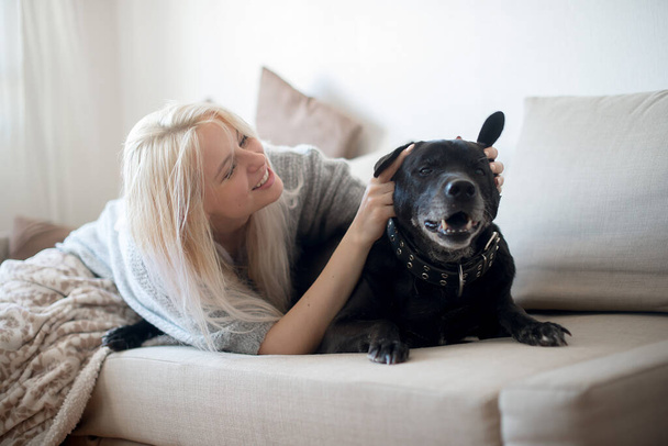 Mujer rubia joven y lindo perro negro en el sofá en casa - Foto, imagen