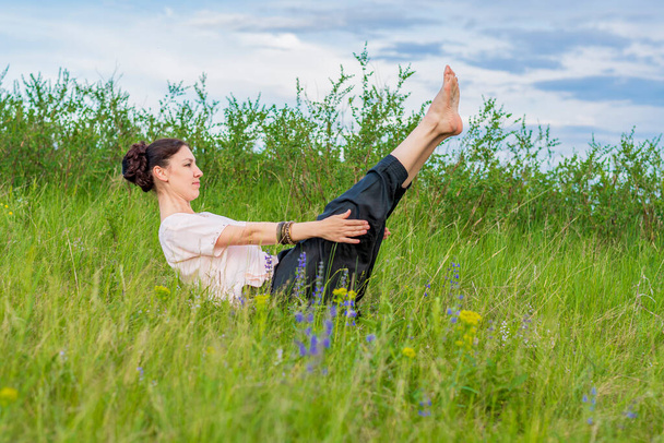 Mulher bonita pratica ioga ao ar livre. Senta-se no chão com as pernas levantadas e segura um canto. - Foto, Imagem