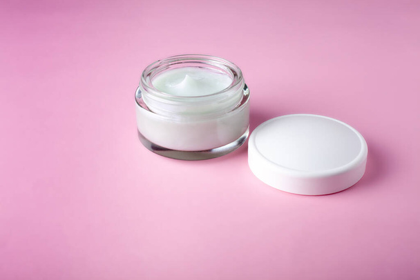 Glass jar and white cap with body cream on pink background. Transparent jar with face cream isolated. Cosmetic product for skin care. - Foto, Imagen