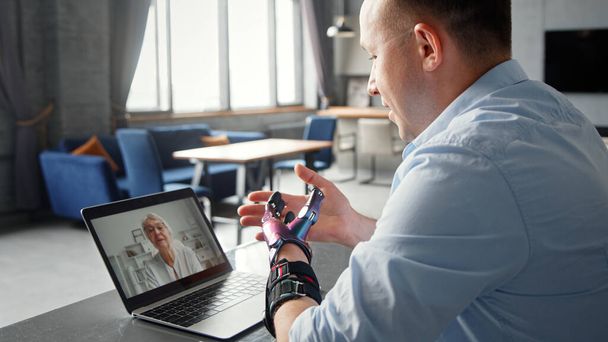 Smiling man with black bio hand prothesis gesticulates talking at online video call - 写真・画像