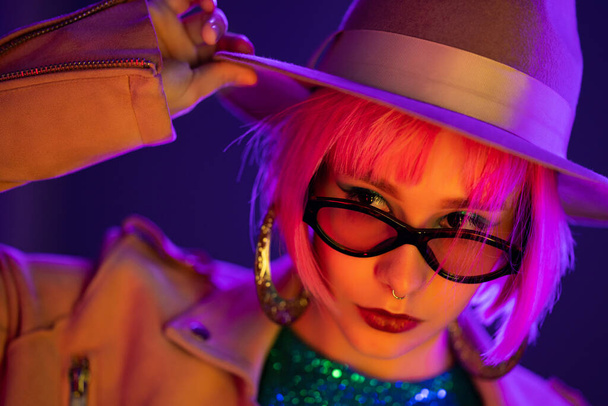 Eccentric woman with pink hair posing under neon light. Charming unusual girl in hat and sunglasses, nightlife concept. Modern model pop outfit, influencer lifestyle. - Fotoğraf, Görsel