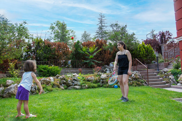 Teenage girl and her baby sister play with tennis racket and ball in the garden - Φωτογραφία, εικόνα