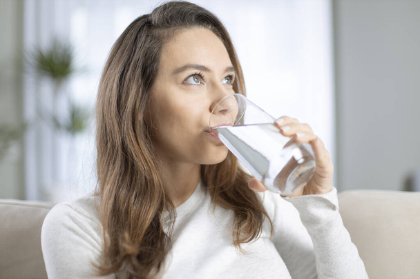 Young woman drinking water sitting on a couch at home. Health benefits of drinking enough water concepts.  - Foto, Imagen