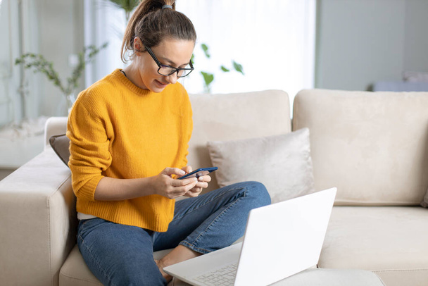 Young woman using mobile phone while sitting a sofa at home  - Fotoğraf, Görsel
