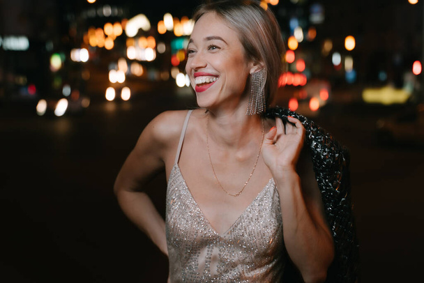 Happy blonde woman looking side. Wide teeth smile with red lips. fancy golden cocktail Glided  dress and long earrings. Big city night life. Bokeh lights on background. Celebration party time mood - Fotó, kép