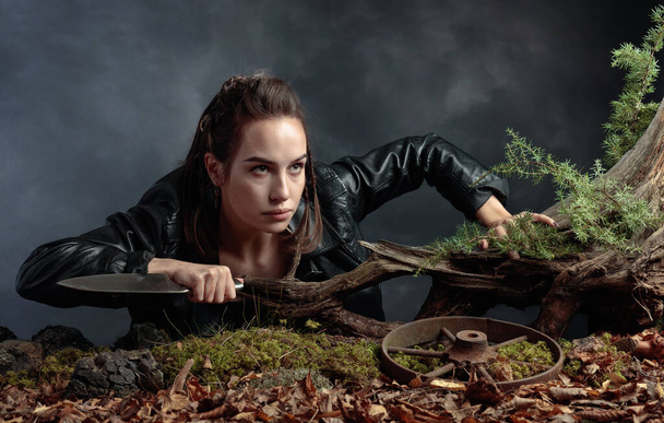 Portrait of a young woman with a knife. A serious girl in a black leather jacket with a knife climbs out of the pit in the forest. Copy space. - Foto, afbeelding