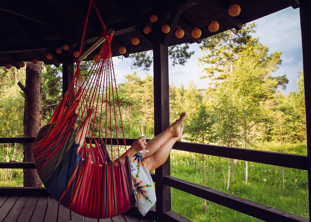 Young woman enjoy alone time in forest resort in Nordic pine tree forest in summer, relax sitting in swing hanging textile chair on balcony, drinking mulled wine. - Fotoğraf, Görsel