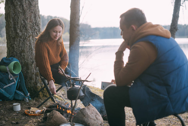 Family couple is resting on the shore of the lake with a tent. Fashing camp with a fire in the forest. A man and woman are shaking silence by fire with hot drink circles - Photo, Image