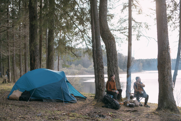 Family couple is resting on the shore of the lake with a tent. Fashing camp with a fire in the forest. A man and woman are shaking silence by fire with hot drink circles - Foto, immagini