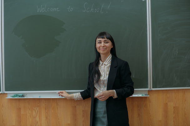 Egy fiatal tanár portréja az iskola tábláján. Gyönyörű tanárnő az osztályteremben a táblánál elmagyarázza a leckét a gyerekeknek. - Fotó, kép