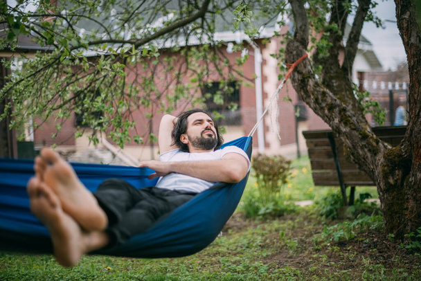 Un hombre descansa en una hamaca en una casa de campo. Un joven yace en una hamaca, dormitando al aire libre en el verano en el verde de los árboles en el campo - Foto, Imagen