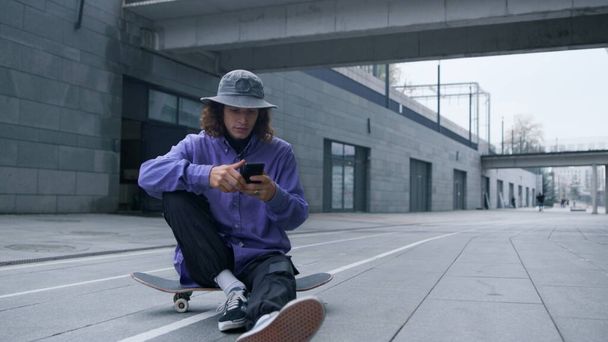 Skater using smartphone outdoor. Hipster sitting on skateboard with phone. - Photo, Image