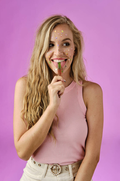 hübsches blondes Mädchen mit Aufklebern im Gesicht leckt ein Bonbon auf rosa Hintergrund - Foto, Bild