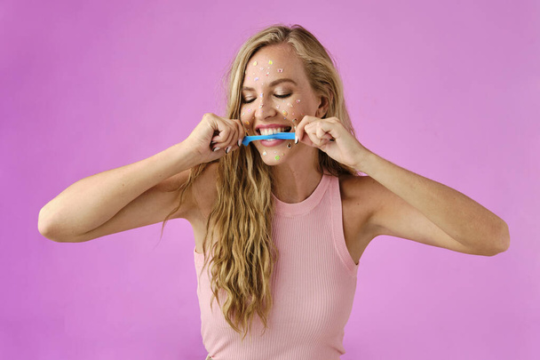 Pretty blonde girl with stickers on her face biting a candy on a pink background - Fotó, kép